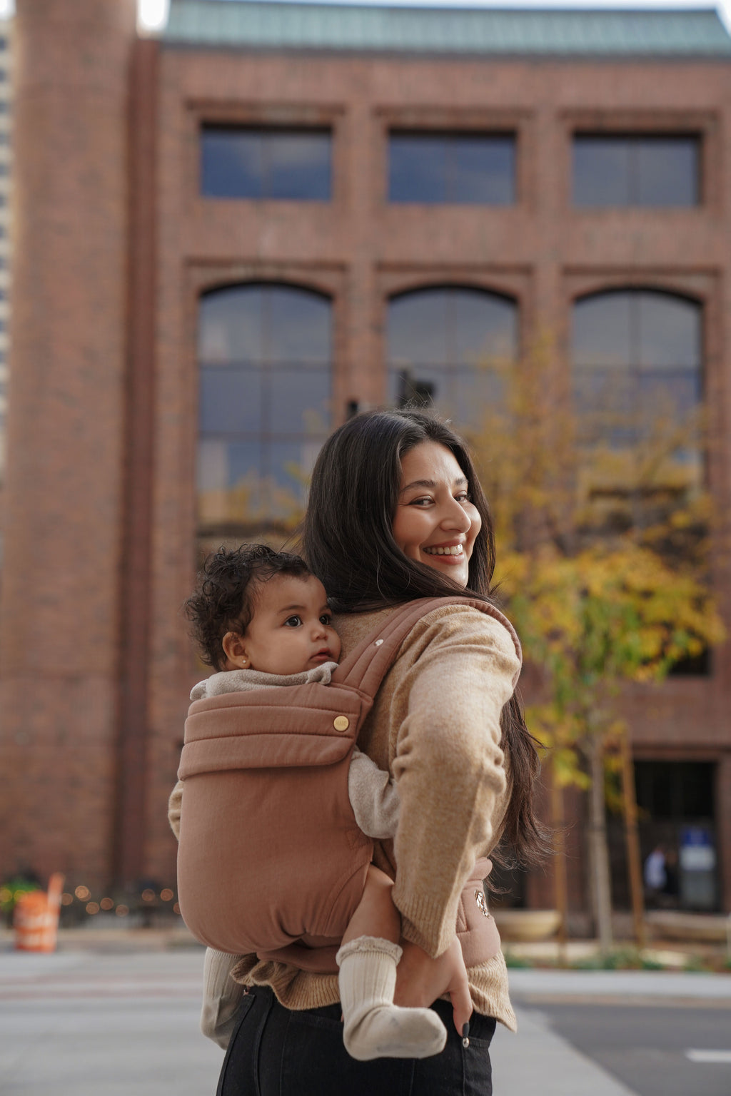 A mom walks through the city wearing her baby on her back in the Mabē Monarch Baby Carrier in Mocha, back carry.