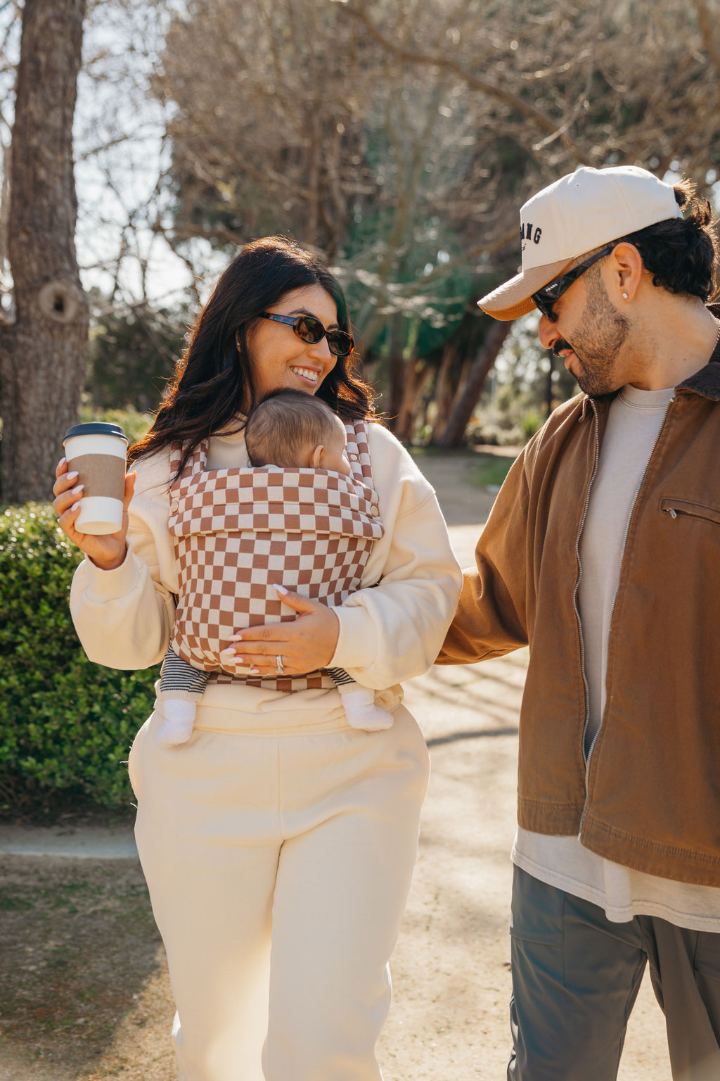 A couple walks outside with coffee while mom wears the baby in the Mabē Monarch Baby Carrier in Mocha Check.