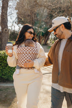 A couple walks outside with coffee while mom wears the baby in the Mabē Monarch Baby Carrier in Mocha Check.
