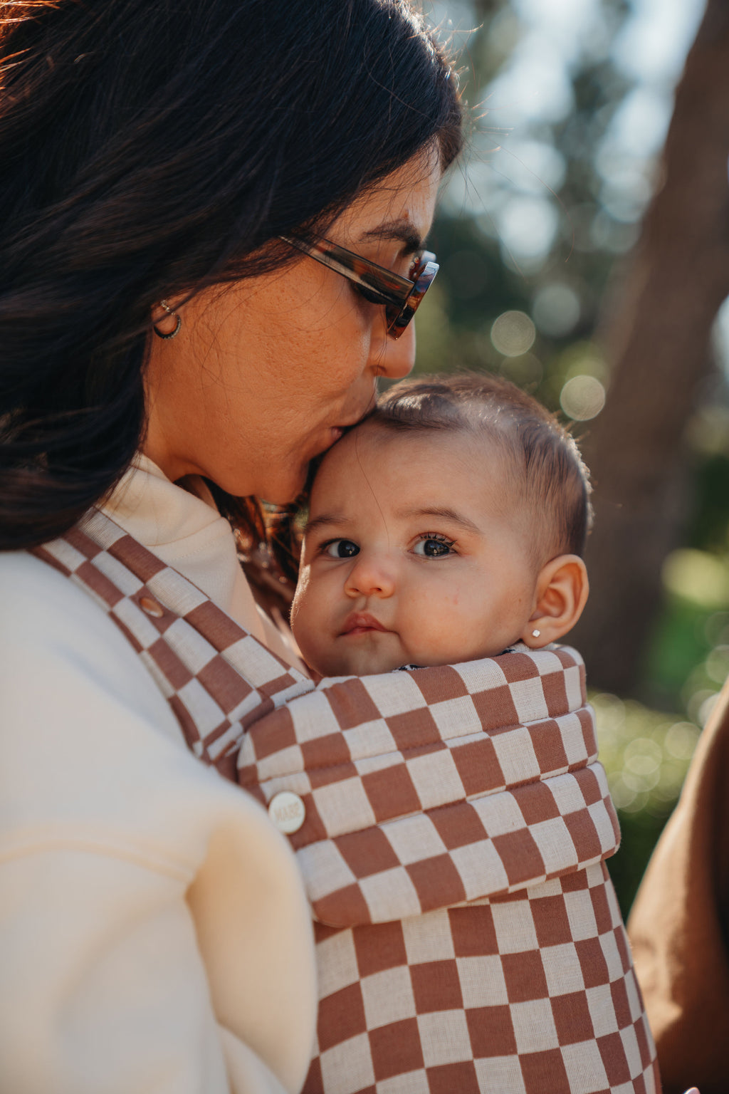 A mom kisses her baby's head while wearing him in the Mabē Monarch Baby Carrier in Mocha Check, close-up of the brown and ecru checkerboard print.