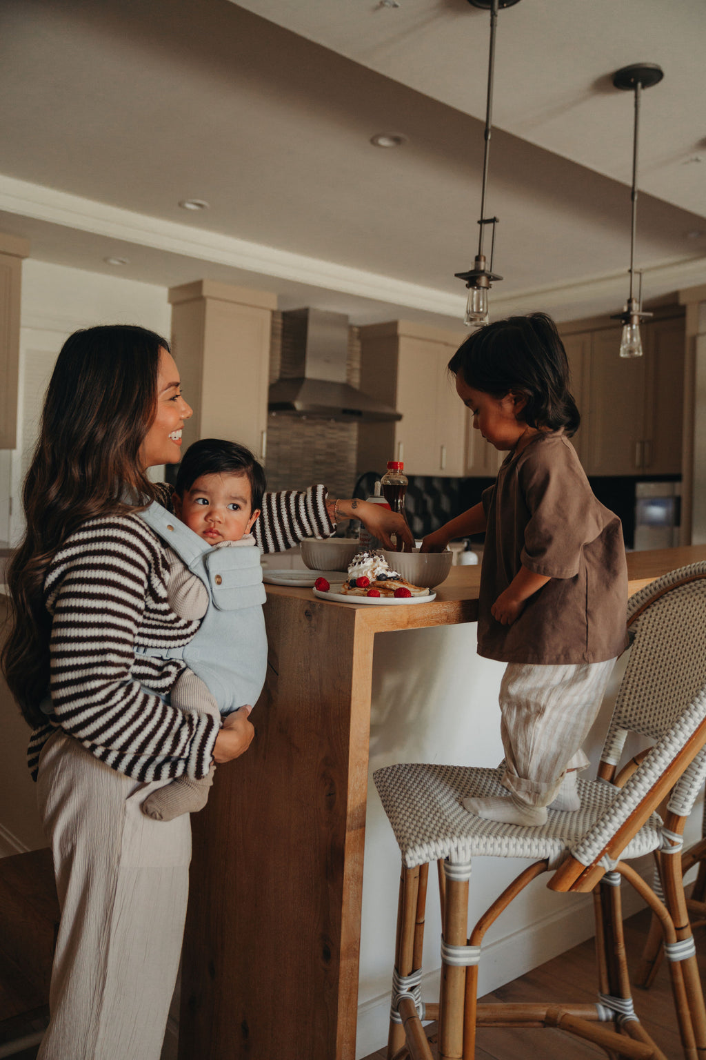 A mom wears her baby in the Mabē Monarch Baby Carrier in Dream Wash Denim in the kitchen while her older child sits at the counter.