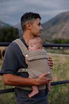 A dad stands by a fence with mountains behind him, wearing his baby in the Mabē Monarch Baby Carrier in Stone Sage.