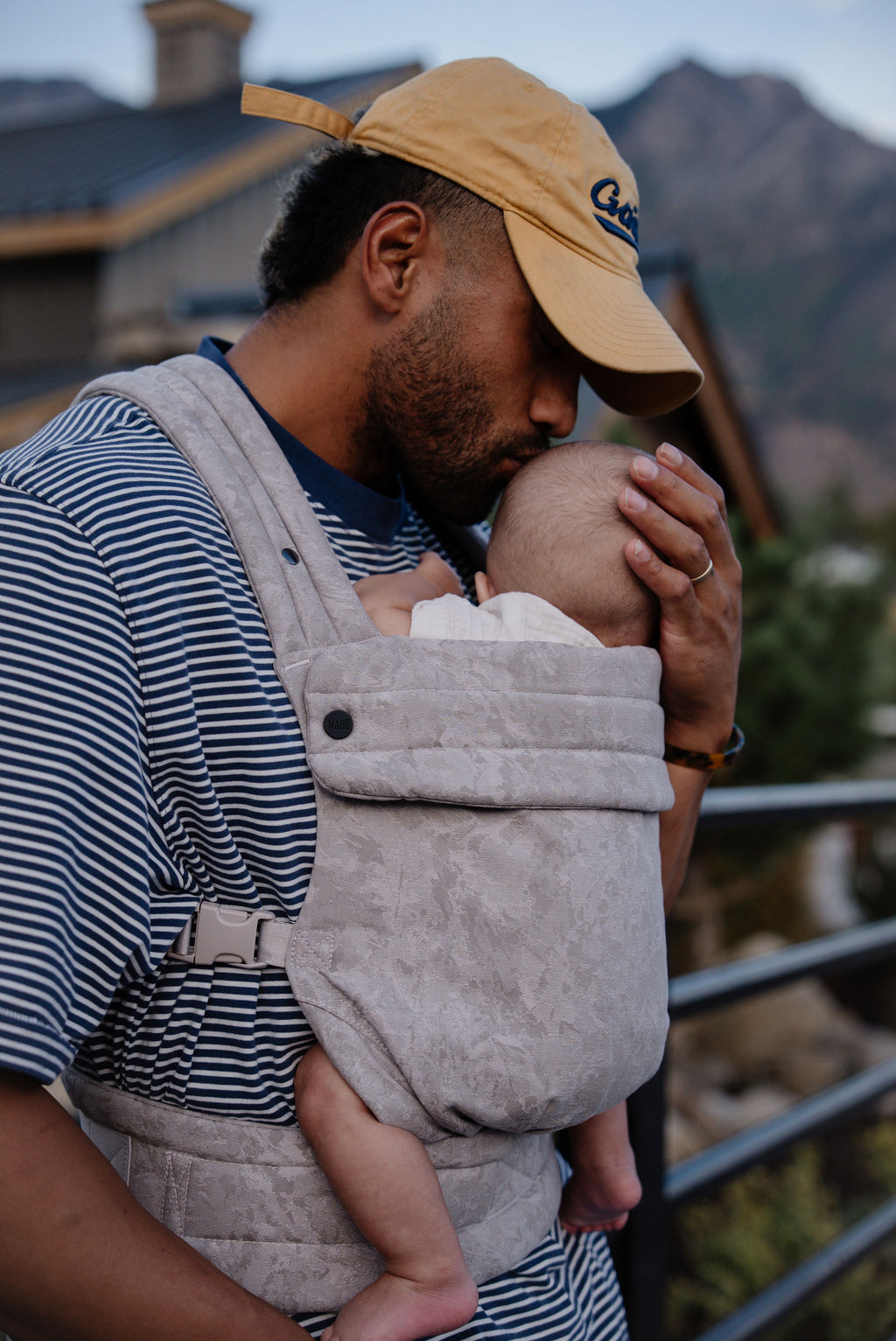 A dad in a cap kisses his baby while wearing him in the Mabē Monarch Baby Carrier in Ashland Camo.