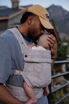 A dad in a cap kisses his baby while wearing him in the Mabē Monarch Baby Carrier in Ashland Camo.