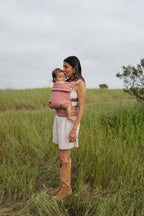 A mom stands in a grassy field wearing her baby in the Mabē Monarch Baby Carrier in Mauve Wings, full length.