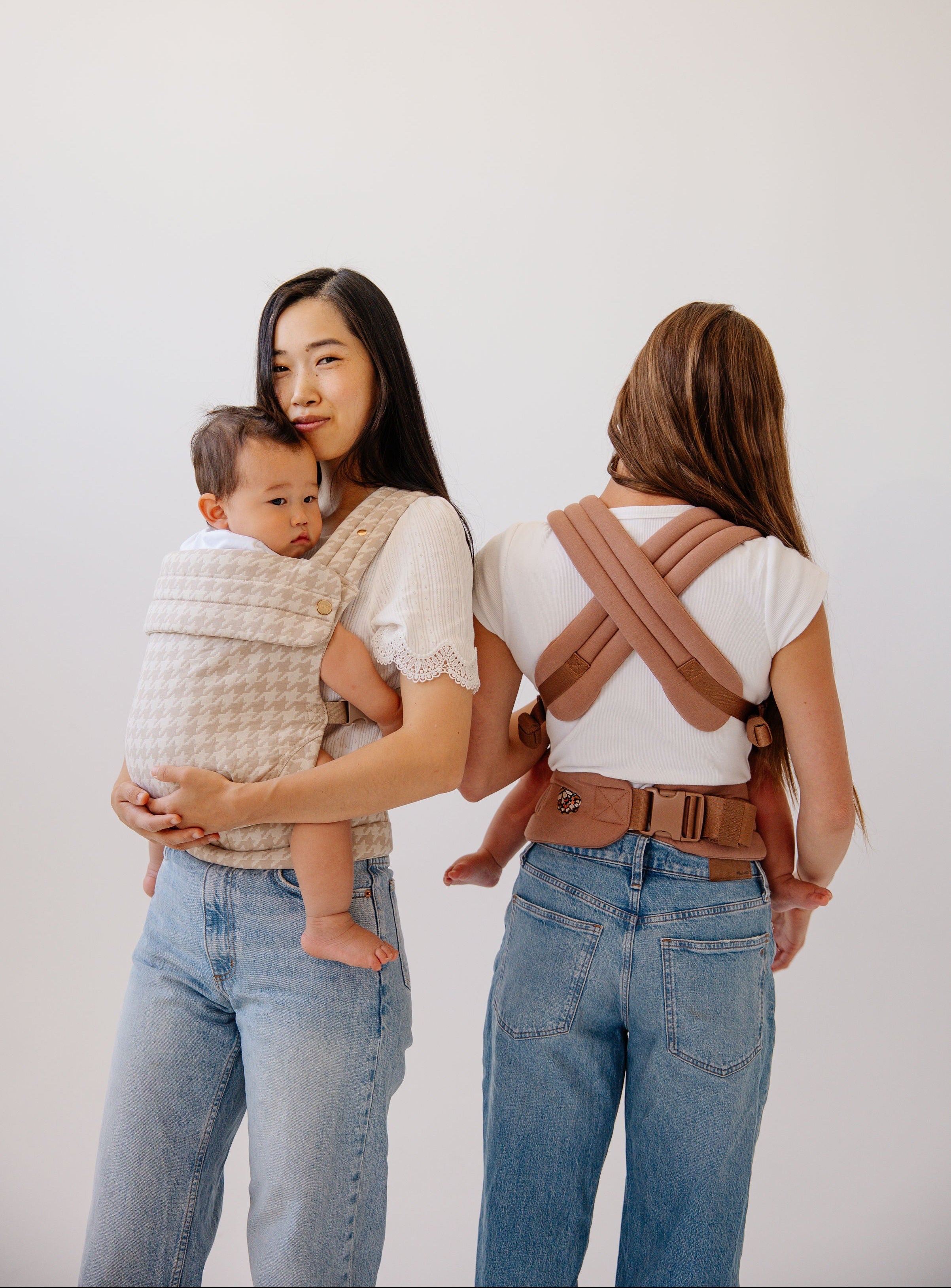 Two women in white shirts and blue jeans holding their babies while wearing a Mabē Buckle Carrier. 