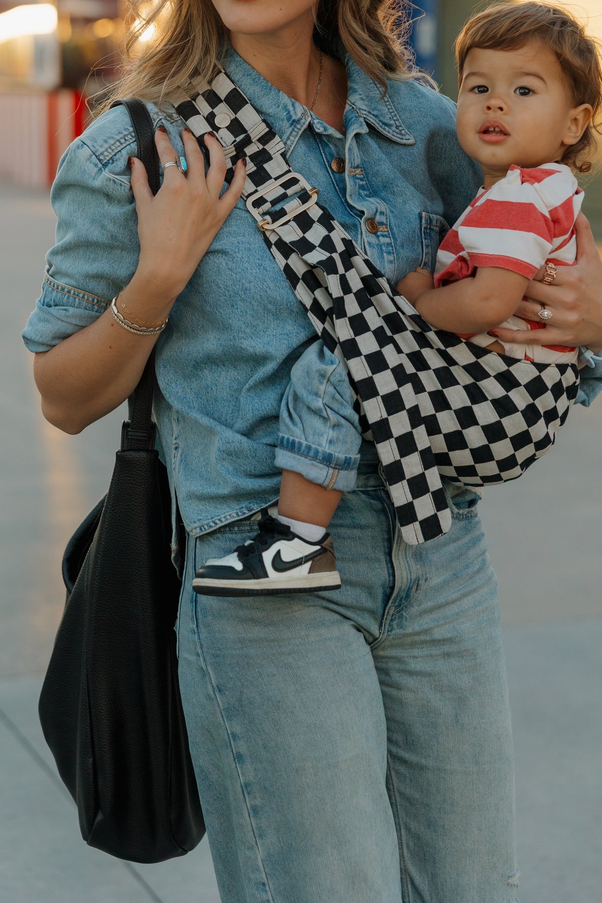 A mom in denim carries her toddler on her hip in the Mabē Monarch Toddler Sling in Ebony Check, close-up of the black and ecru check.