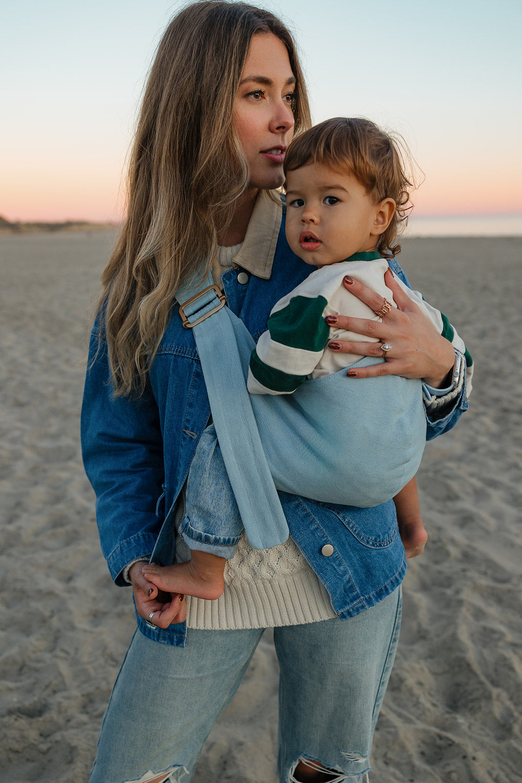 Woman with long, curled, highlighted brown hair holding a child with light brown curly hair in a Mabē toddler sling in Dream Wash Denim on a sandy beach at sunset. The child is wearing a white sweatshirt with dark green thick stripes on the sleeves. The woman wears a medium wash denim jacket and light wash ripped denim jeans. A cable knit cream-colored sweater peeks out from under the child and jacket.