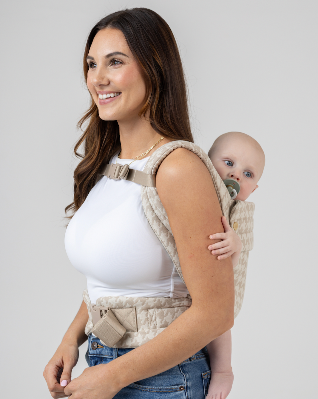 A mom wears her baby in a back carry with the Mabē Monarch Carrier Chest Clip fastened across the shoulder straps, side view.