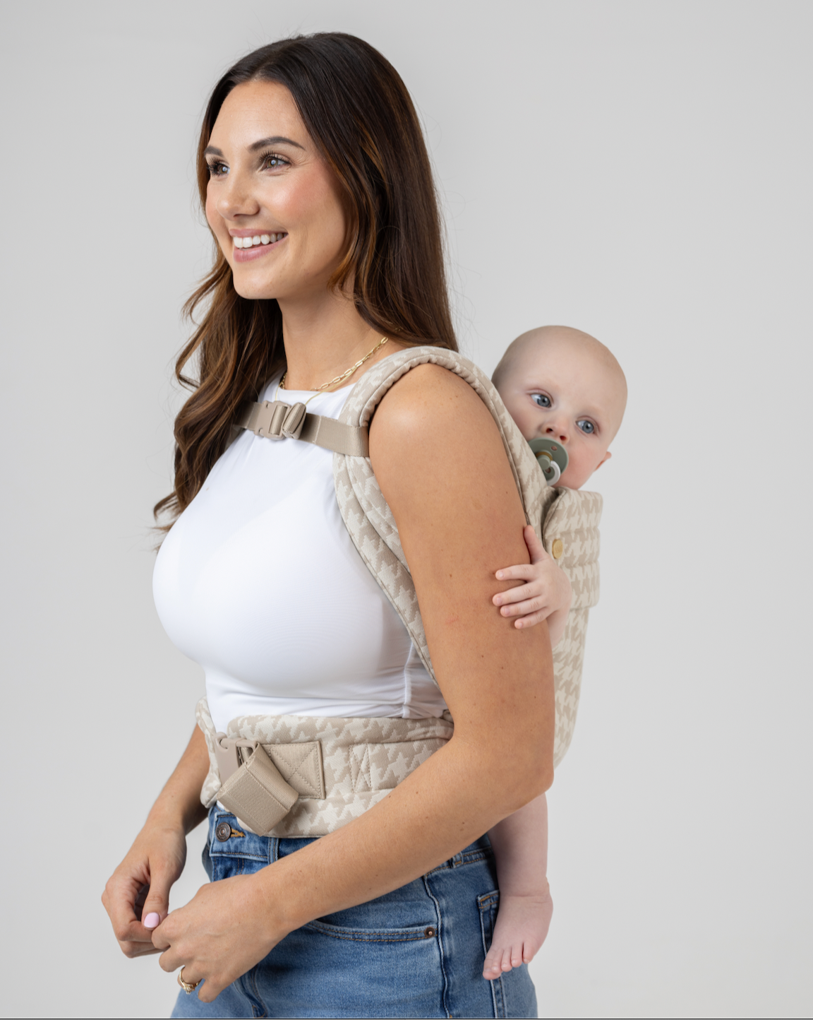 A mom wears her baby in a back carry with the Mabē Monarch Carrier Chest Clip fastened across the shoulder straps, side view.