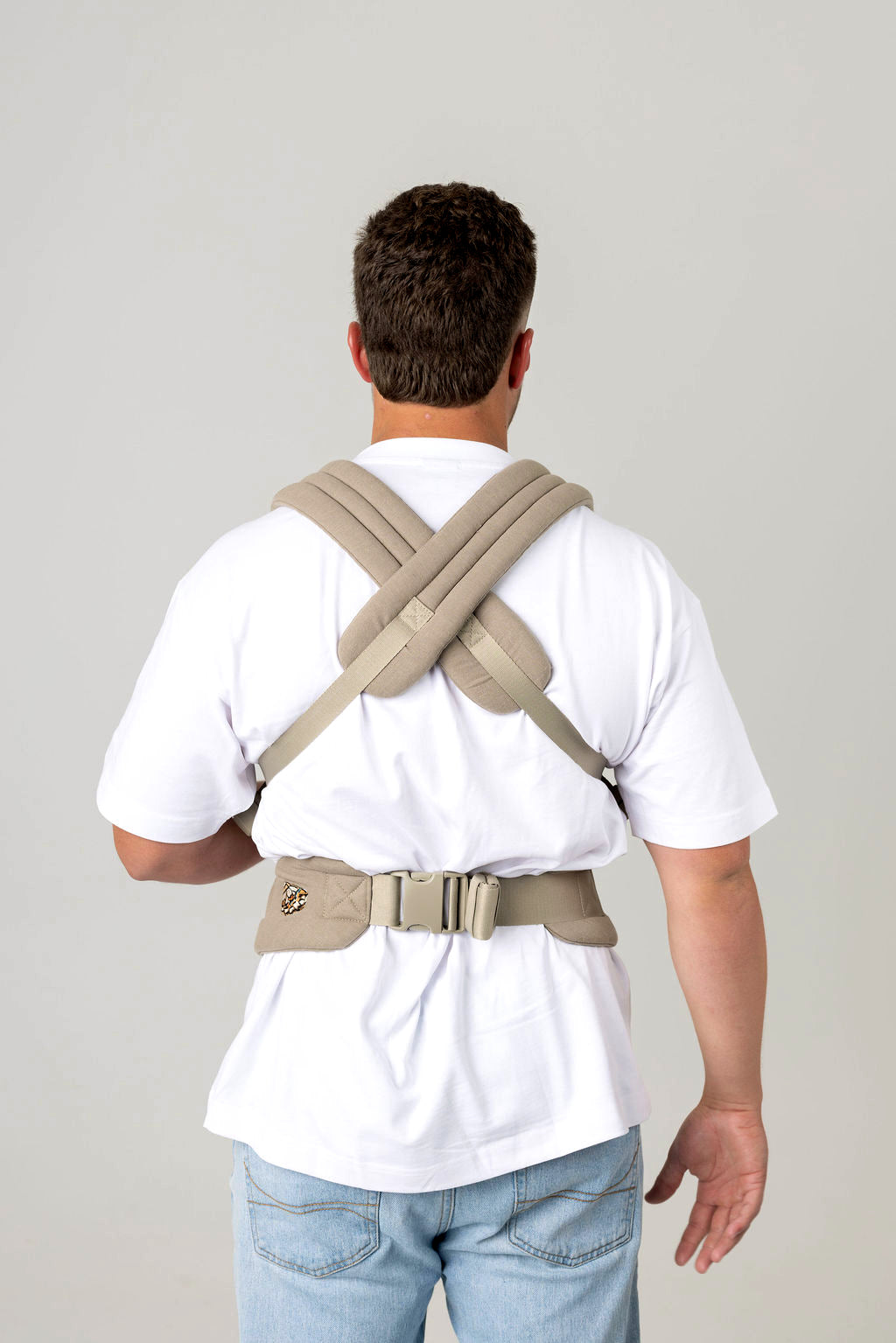 Waist-to-above-head photo of a white man with brown hair from the back wearing a greenish-gray baby carrier with the straps crossed across his back and the waistband buckled around his bag, against a light background. He is wearing a white t-shirt and light wash jeans which are only just visible.