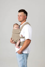 Waist-to-above-head photo of a white man with brown hair cut in a mullet with a fade and slight facial hair, carrying a baby in a greenish-gray carrier against a light background. He is wearing a white t-shirt and light wash jeans which are only just visible.