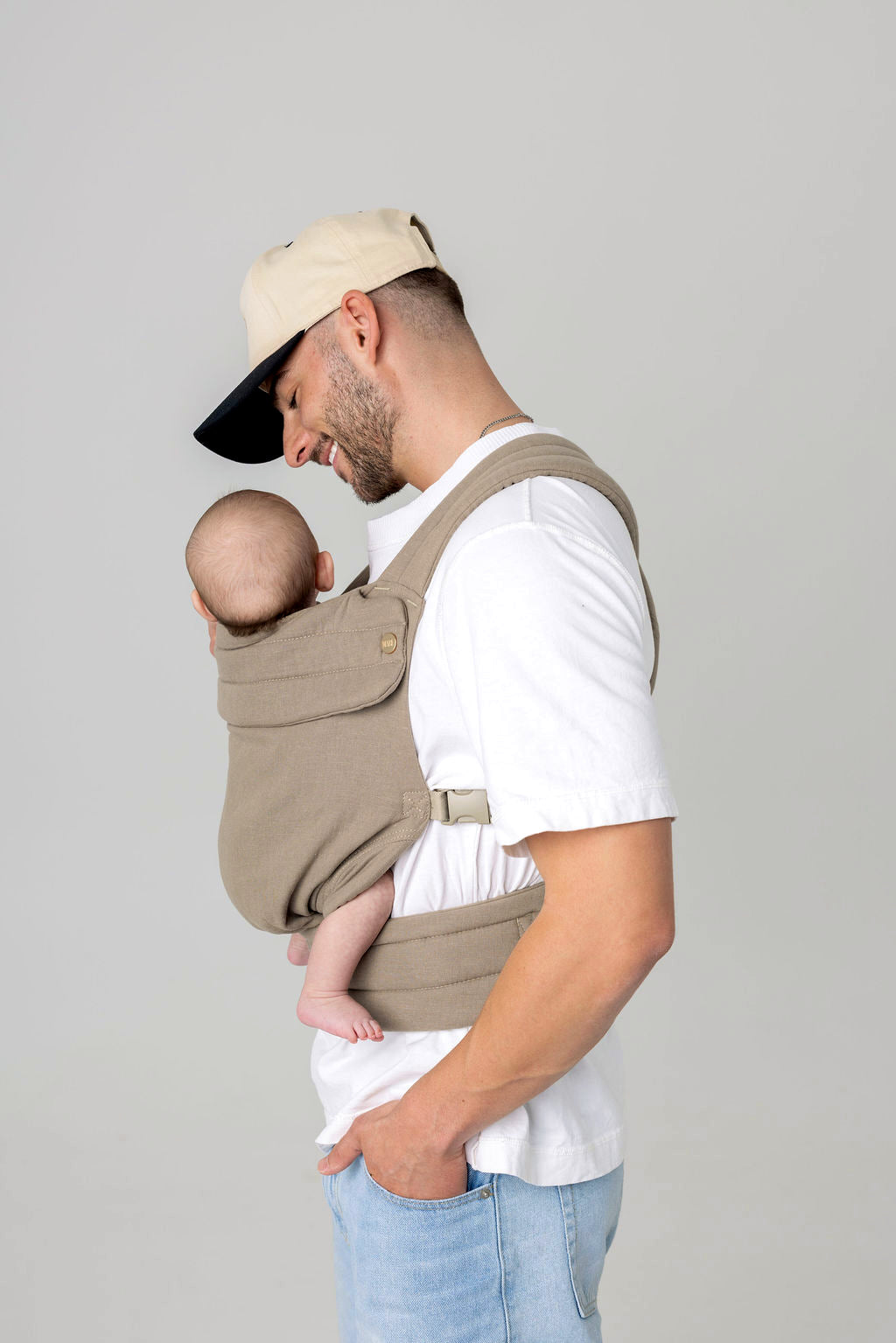 Waist-to-above-head photo of a white man with dark facial hair and a light-colored hat with a dark brim, carrying a baby in a greenish-gray carrier against a light background. He is wearing a white t-shirt and light wash jeans which are only just visible.
