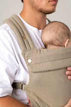 Cropped photo of a white man with dark facial hair carrying a baby with dark wispy hair in a greenish-gray carrier against a light background. He is wearing a white t-shirt. The baby is looking away from the camera.