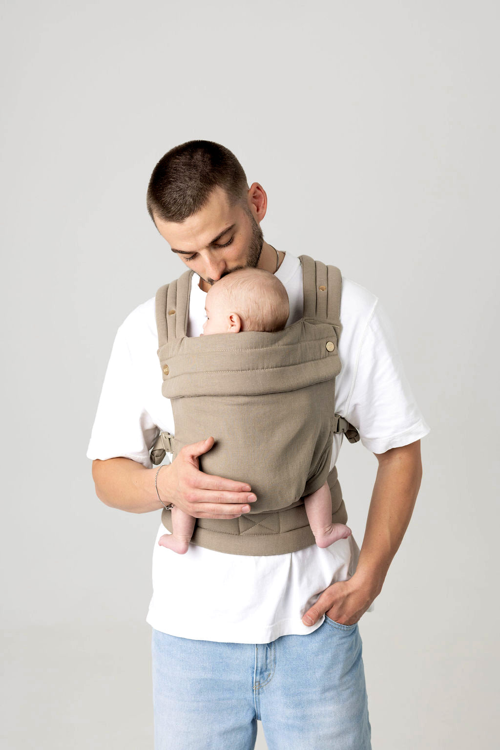 Waist-to-above-head photo of a white man with dark facial hair carrying a baby in a greenish-gray carrier against a light background. He is wearing a white t-shirt and light wash jeans which are only just visible.