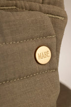Close-up of a solid, greenish-stone-colored fabric baby carrier head support with a gold snap with the 'MABE' logo. Matching stitching is visible. The background is white.