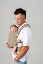 Waist-to-above-head photo of a white man with brown hair cut in a mullet with a fade and slight facial hair, carrying a baby in a greenish-gray carrier against a light background. He is wearing a white t-shirt and light wash jeans which are only just visible.