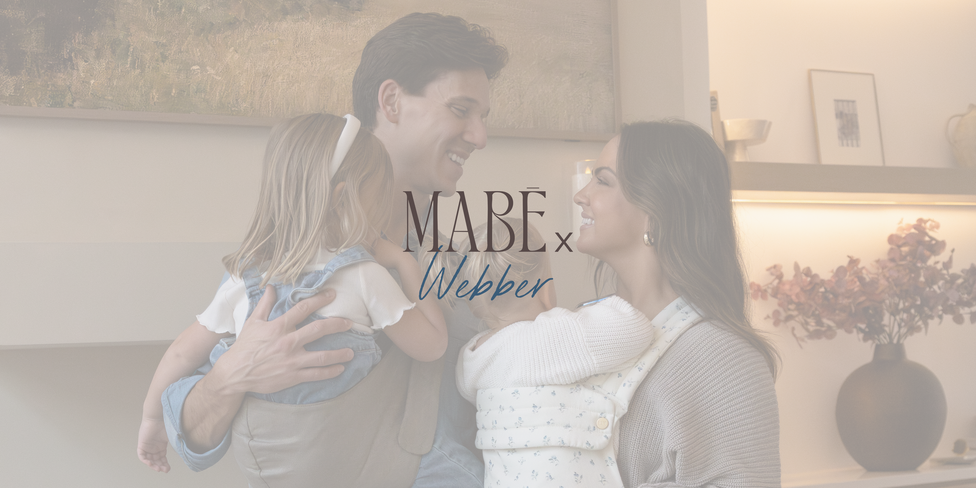 The Webber family of four in a cozy living room wearing Mabē Monarch Buckle Carrier and Toddler Sling with 'The Mabe x Webber Collection' text overlay.