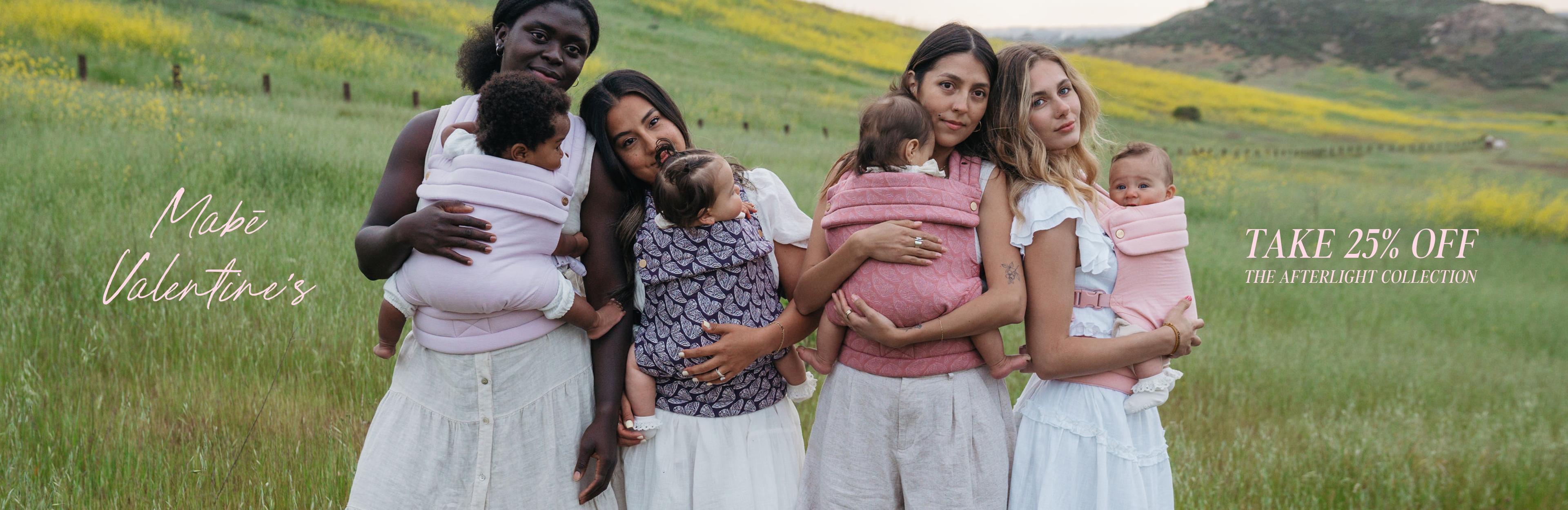 Four women in a field with wildflowers and a fence. They all wear white clothing and babies in Mabē Monarch buckle carriers in solid lilac, dark purple with butterfly wings, medium pink with butterfly wings, and solid light pink. The first woman has dark skin and black hair. The second and third have straight dark hair and tan skin. The fourth has brown and blonde hair, light skin. Text reads Mabē Valentine's, TAKE 25% OFF THE AFTERLIGHT COLLECTION.