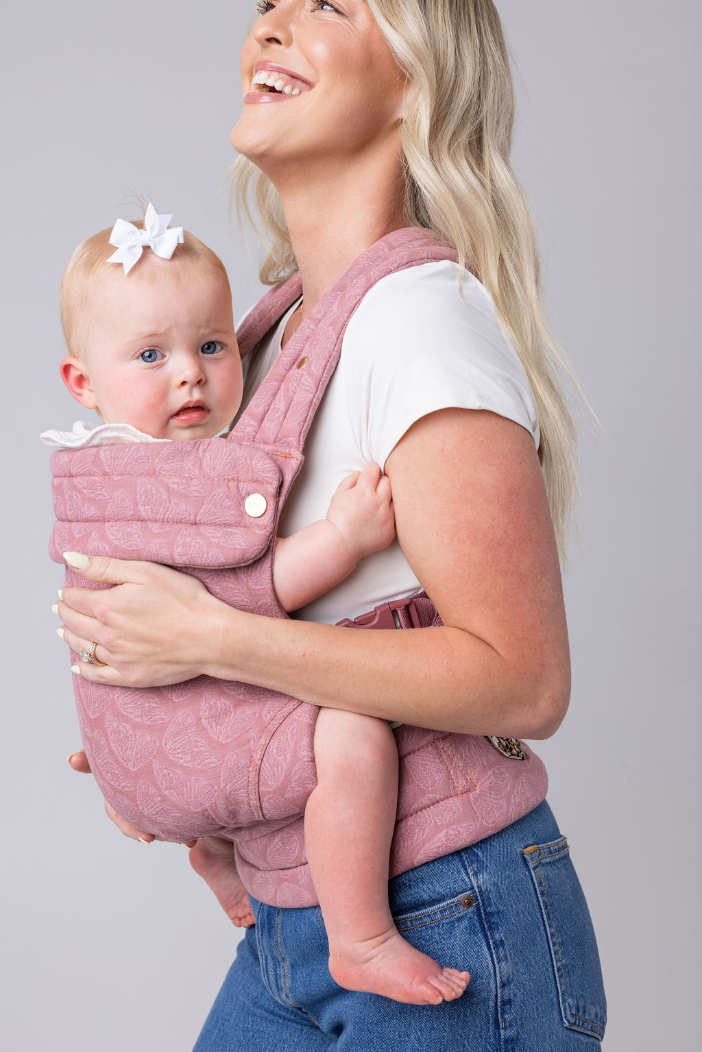 A closed-in photo of a light-skinned woman with long blonde hair, a white t-shirt, and medium-wash jeans. She carries a light-skinned, light-haired baby in the Mabē Monarch Buckle carrier in Mauve Wings, which is blush pink with lighter butterfly wings patterned on it. She faces the left of the frame and is shown from the hips up and eyes down. The background is white or light gray.