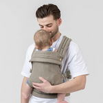 The Mabē Monarch Buckle Carrier in Stone Sage, which is a solid soft greeny stone color cotton/linen fabric, worn by a man with a baby on his front.