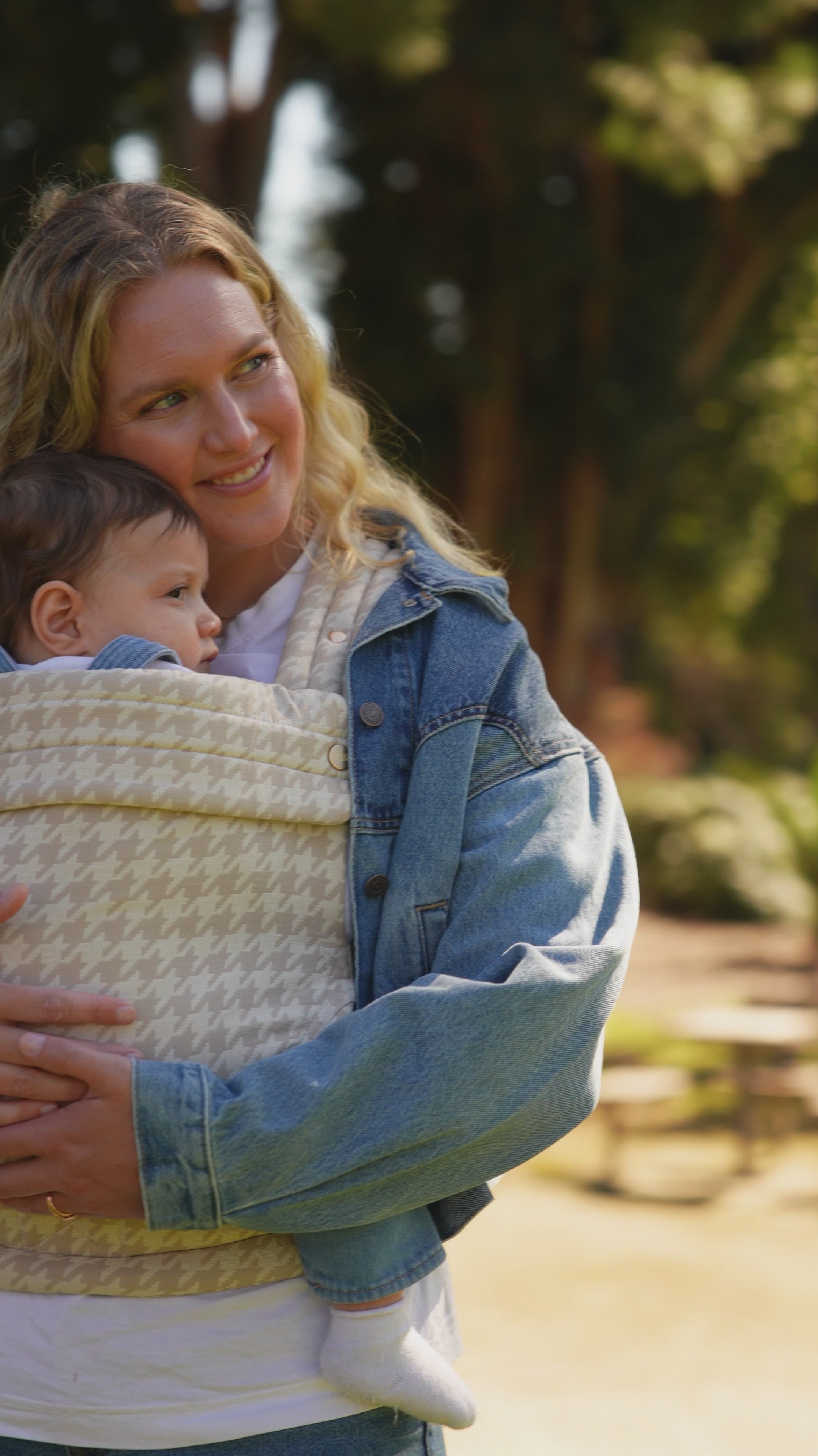 Video of a mom in a denim jacket wearing her baby in the Mabē Monarch Baby Carrier in Houndstooth outdoors.