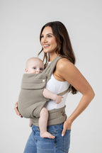 A woman with long brown hair with highlights and tanned skin wears a white tank top and the Mabē Monarch Buckle Carrier in Stone Sage, which is a solid, green-tinged gray color. The carrier holds a light-skinned baby with blue eyes. She wears medium wash jeans and faces the left of the frame with a big smile. The baby is looking at the viewer. His arms and legs are bare.