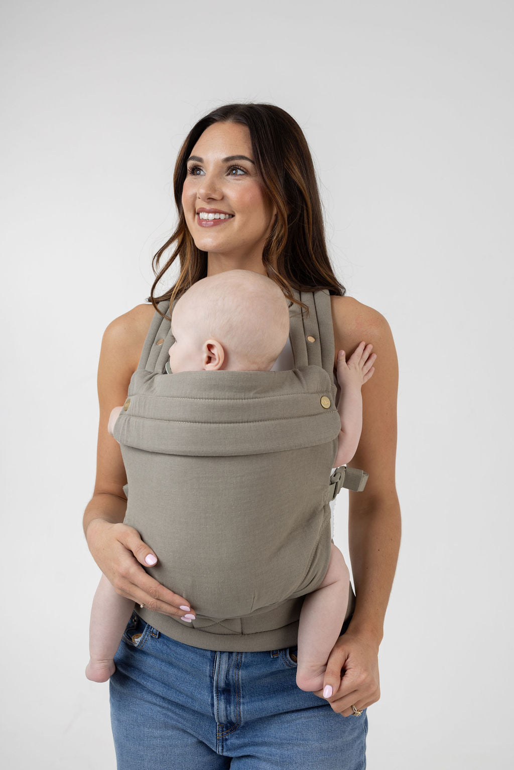 A woman with long brown hair with highlights and tanned skin wears a white tank top and the Mabē Monarch Buckle Carrier in Stone Sage, which is a solid, green-tinged gray color. The carrier holds a light-skinned baby with blue eyes. She wears medium wash jeans and faces the viewer but looks to the left of the frame with a big smile. The baby is looking to his left also. His arms and legs are bare.