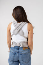 An unseen person with long brown hair with highlights and tanned skin is shown from behind. They wear a white tank top and the Mabē Monarch Buckle Carrier in Ashland Camo, which is a light gray camouflage pattern. Only the carrier's straps crossed across the back and the waistband are visible. The top part of the person's medium wash jeans are visible but the picture is cropped top of thigh and below.