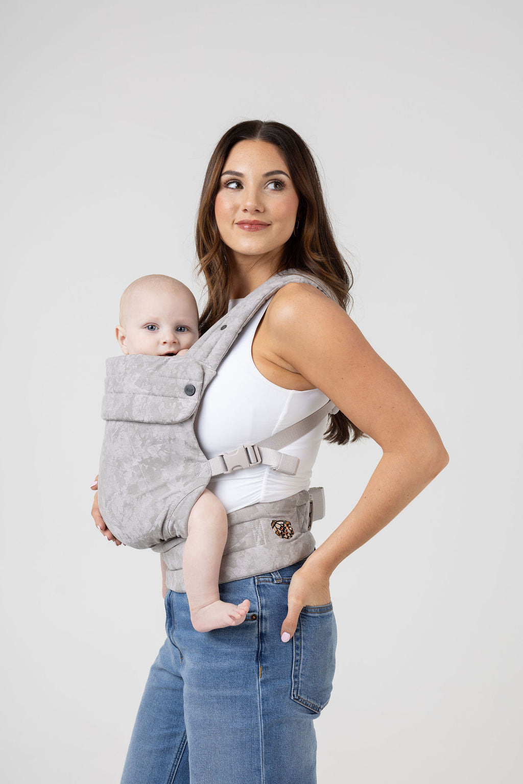 A woman with long brown hair with highlights and tanned skin wears a white tank top and the Mabē Monarch Buckle Carrier in Ashland Camo, which is a light gray camouflage pattern. The carrier holds a light-skinned baby with blue eyes. She wears medium wash jeans and faces the left of the frame but looks to the right of the frame with a slight smile. The baby is looking at the viewer. His arms and legs are bare.