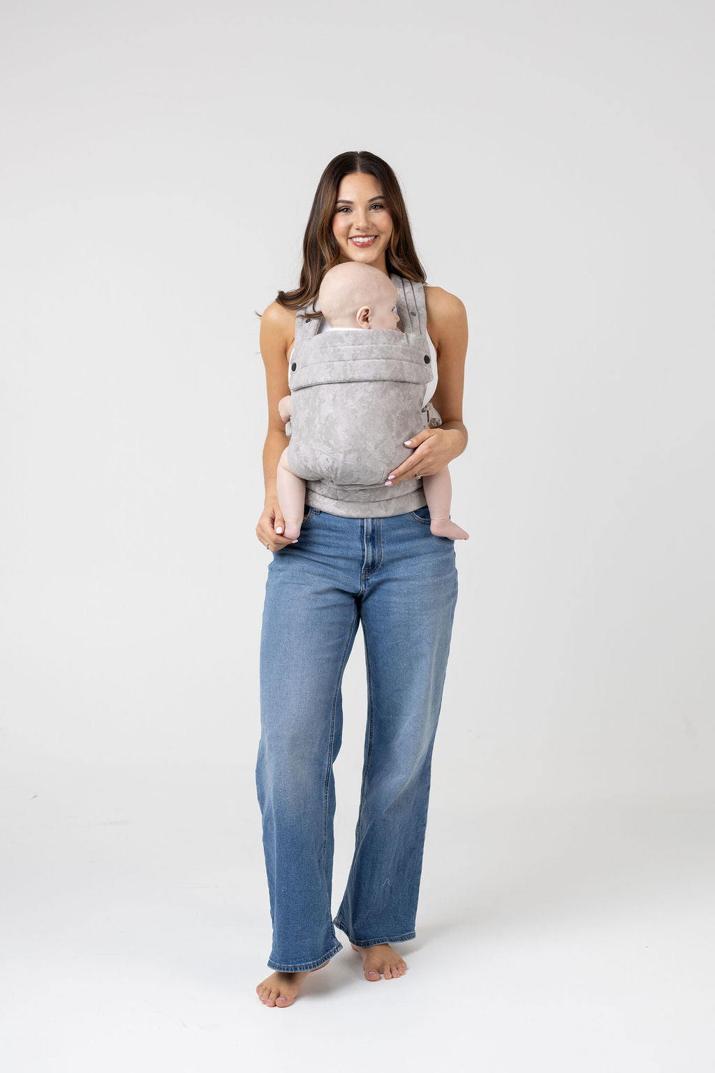 Full-length photo of a woman with long brown hair with highlights and tanned skin wearing a white tank top and the Mabē Monarch Buckle Carrier in Ashland Camo, which is light gray and camouflage patterned. Inside is a light-skinned baby. She wears medium wash wide leg jeans and faces the viewer with a big smile. The baby is looking to the left of the frame. His legs are bare and the woman is barefoot.