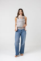 Full-length photo of a woman with long brown hair with highlights and tanned skin wearing a white tank top and the Mabē Monarch Buckle Carrier in Ashland Camo, which is light gray and camouflage patterned. Inside is a light-skinned baby. She wears medium wash wide leg jeans and faces the viewer with a big smile. The baby is looking to the left of the frame. His legs are bare and the woman is barefoot.