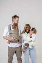 The Mabē Monarch Toddler Sling in  Sunday Floral, a white fabric with tiny sprigs of blue flowers, and the Mabē Monarch Buckle Carrier in Charles Herringbone in shades of brown shown with a family of 4.