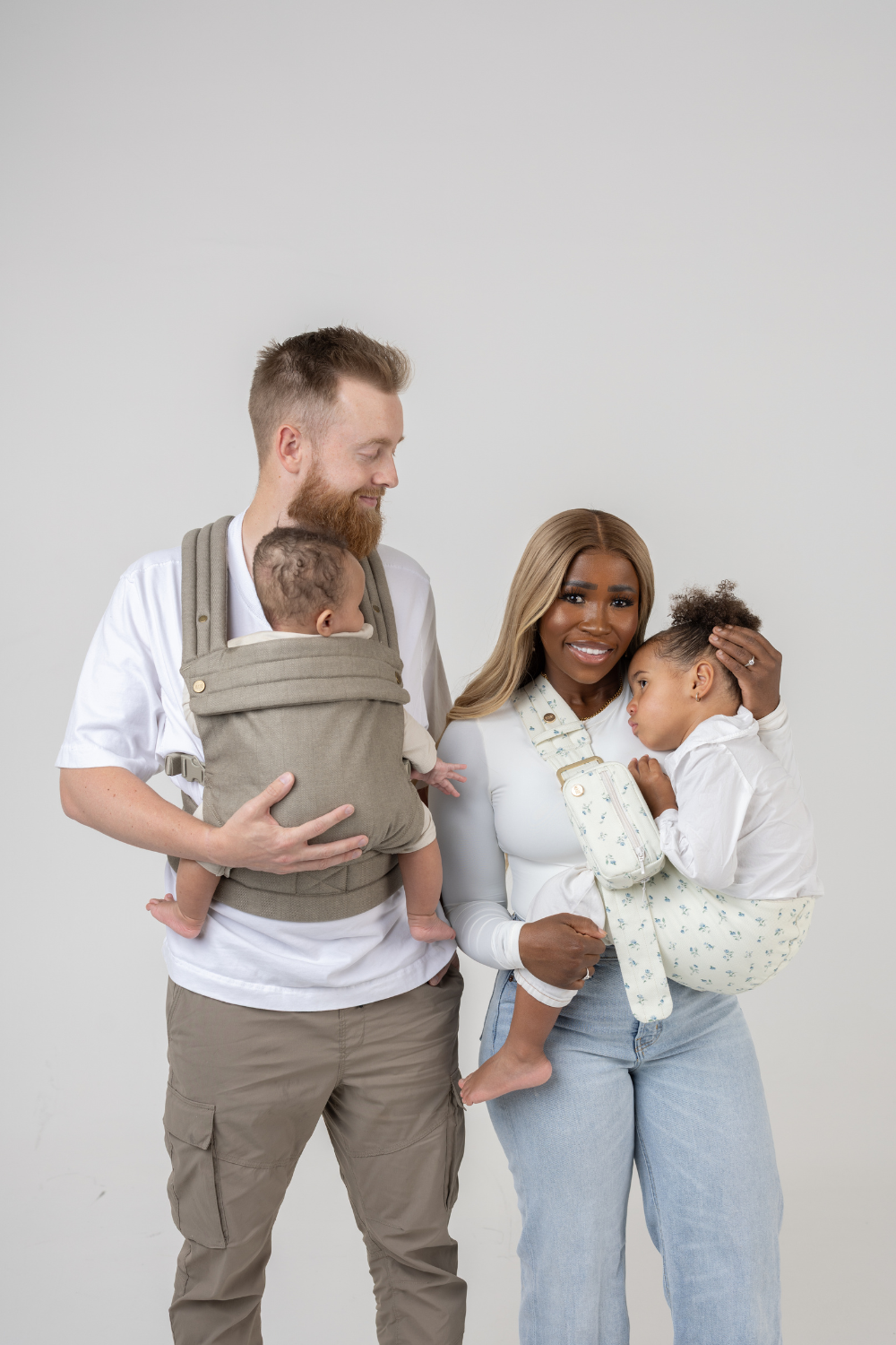 The Mabē Monarch Toddler Sling in  Sunday Floral, a white fabric with tiny sprigs of blue flowers, and the Mabē Monarch Buckle Carrier in Charles Herringbone in shades of brown shown with a family of 4.
