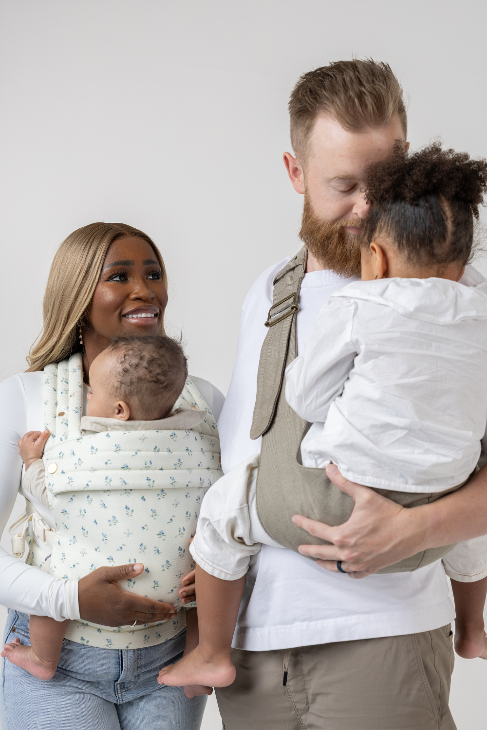 The Mabē Monarch Toddler Sling in Charles Herringbone in shades of brown and the Mabē Monarch Buckle Carrier in Sunday Floral, a white fabric with tiny sprigs of blue flowers, shown with a family of 4.