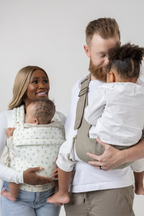 The Mabē Monarch Toddler Sling in Charles Herringbone in shades of brown and the Mabē Monarch Buckle Carrier in Sunday Floral, a white fabric with tiny sprigs of blue flowers, shown with a family of 4.