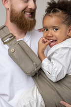 Man wearing the Mabē Monarch Toddler Sling in Charles Herringbone—delicate herringbone weave cotton/linen fabric in shades of brown—to carry a toddler girl.