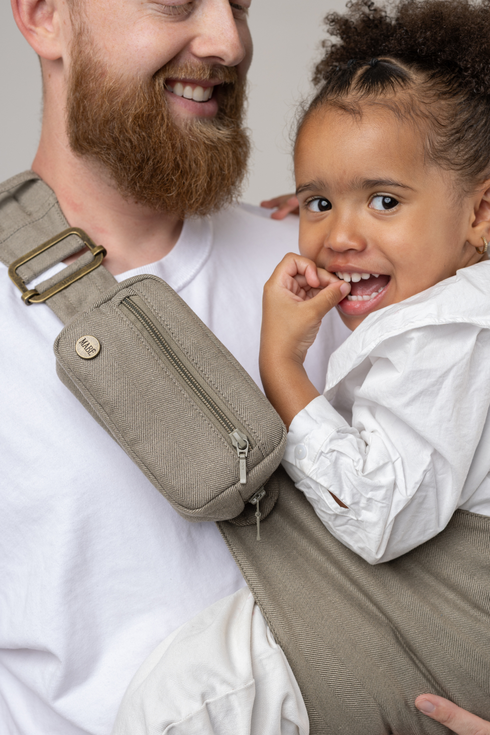 Man wearing the Mabē Monarch Toddler Sling in Charles Herringbone—delicate herringbone weave cotton/linen fabric in shades of brown—to carry a toddler girl.
