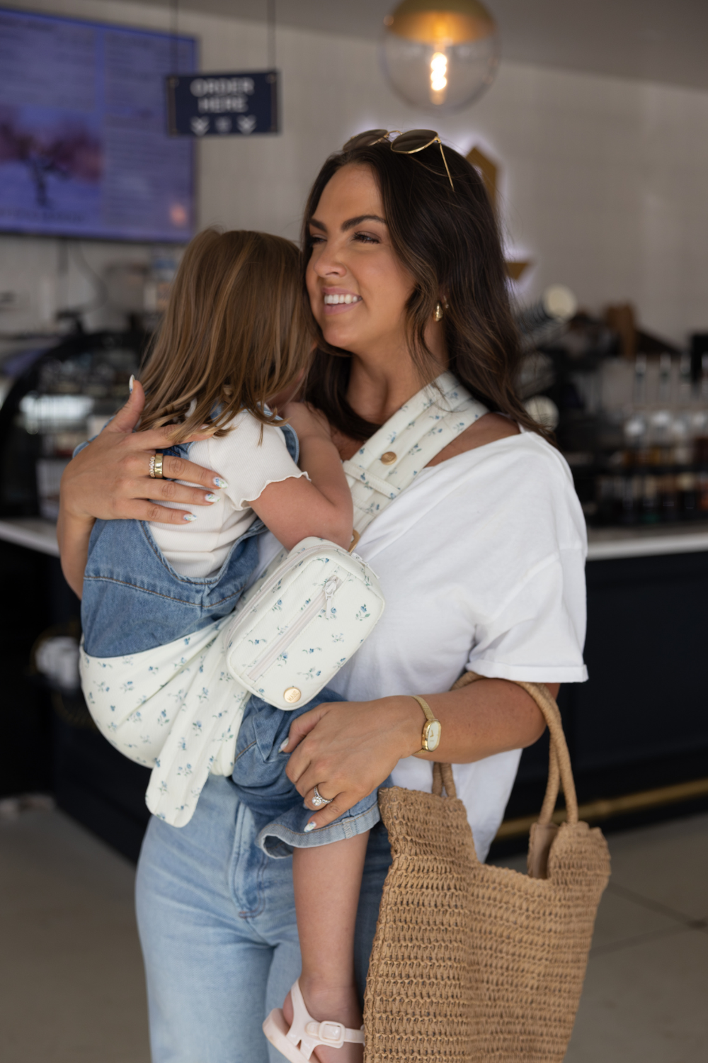 Liz Webber wearing the Mabē Monarch Toddler Sling and Accessory Bag in Sunday Floral—white with tiny sprigs of blue flowers—to carry a toddler girl.
