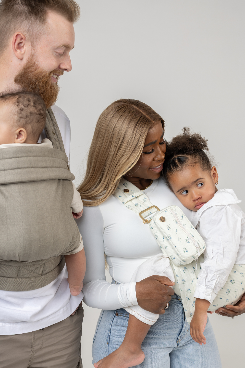 The Mabē Monarch Toddler Sling in  Sunday Floral, a white fabric with tiny sprigs of blue flowers, and the Mabē Monarch Buckle Carrier in Charles Herringbone in shades of brown shown with a family of 4.