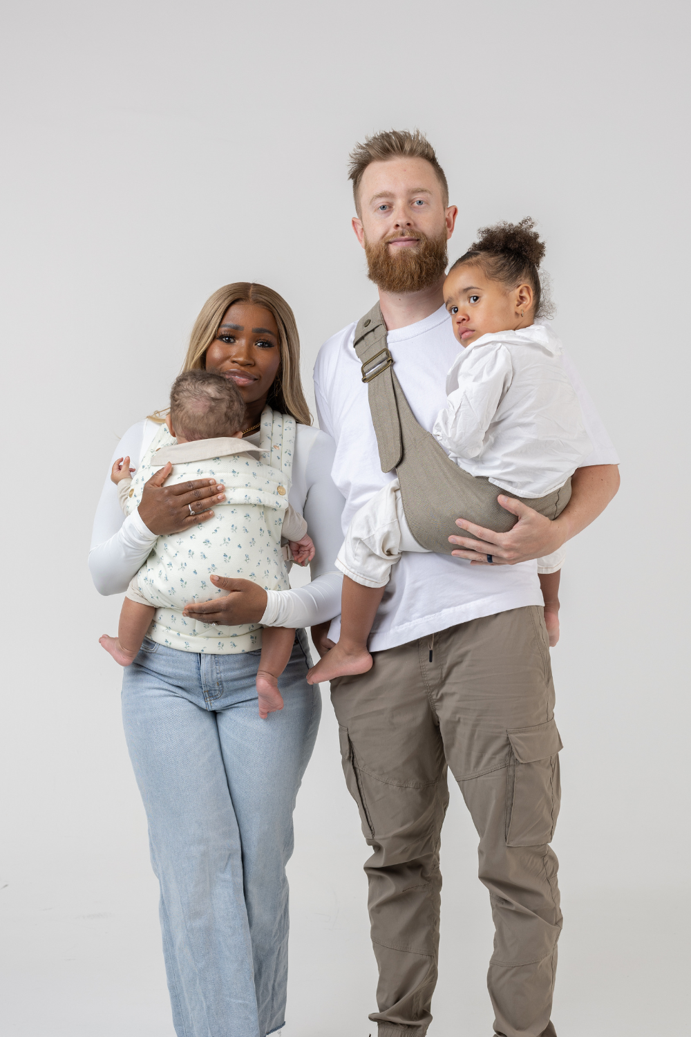 The Mabē Monarch Toddler Sling in Charles Herringbone in shades of brown and the Mabē Monarch Buckle Carrier in Sunday Floral, a white fabric with tiny sprigs of blue flowers, shown with a family of 4.