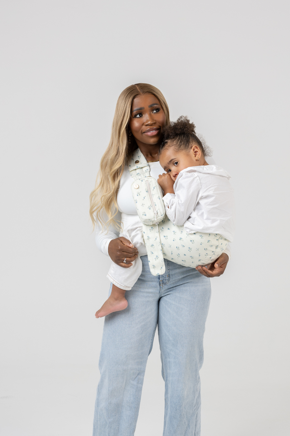 The Mabē Monarch Toddler Sling and Accessory Bag in Sunday Floral, which is solid white with tiny sprigs of blue flowers worn by a woman with a toddler girl.