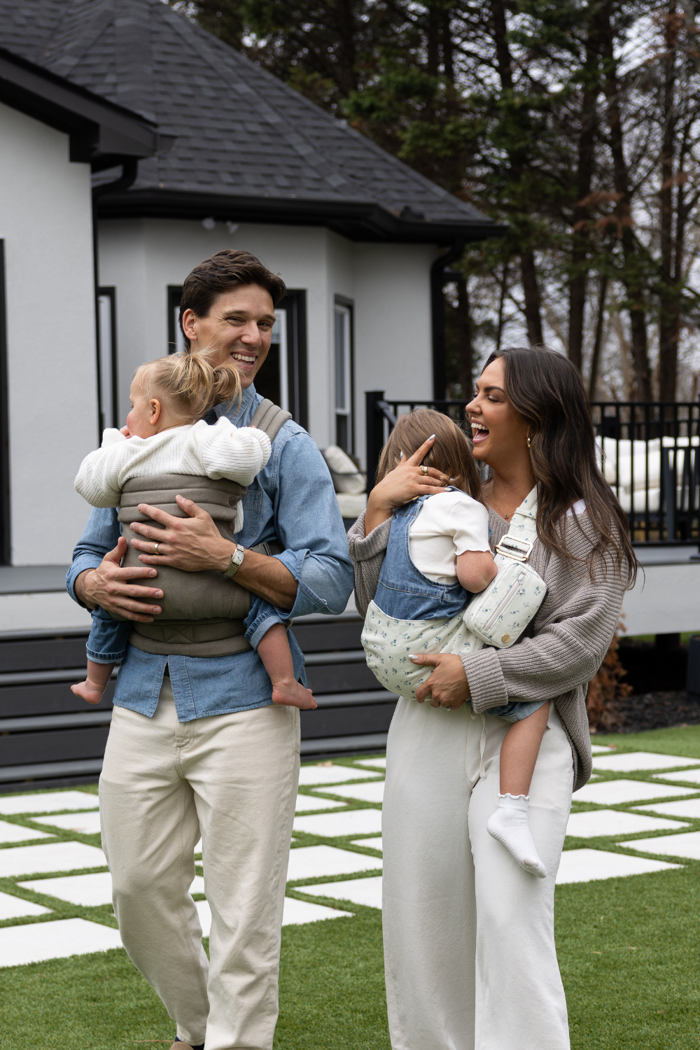 The Mabē Monarch Toddler Sling in Sunday Floral (solid white with tiny sprigs of blue flowers) and Mabē Monarch Buckle Carrier in Charles Herringbone (delicate herringbone pattern in shades of brown) worn by Liz and Zach Webber with their two children.